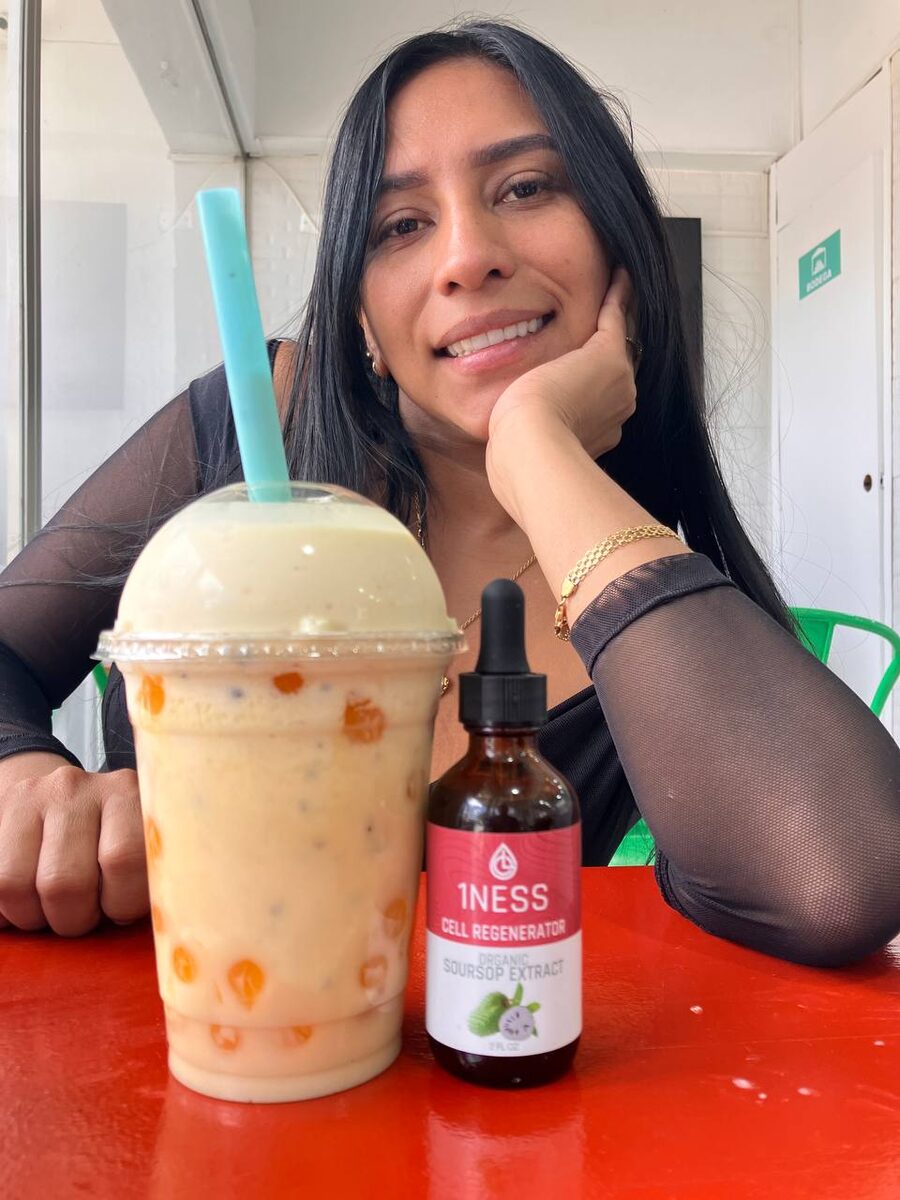 A smiling woman at a cafe table with a soursop smoothie and a 1NESS soursop extract bottle